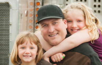 Man with two little girls hugging him in front of semi truck