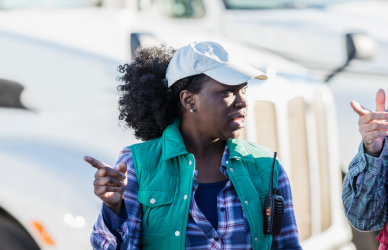 black woman wearing ball cap hat and flannel talking with man holding clipboard in front of parked semi trucks